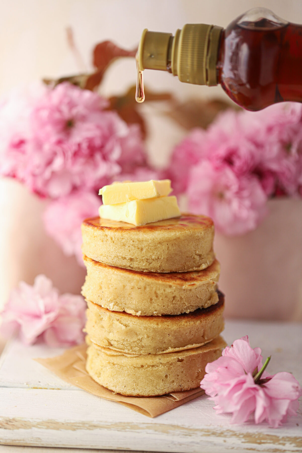 A stack of fluffy pancakes with a block of butter and a drizzle of maple syrup. Pink blossoms are in the background.