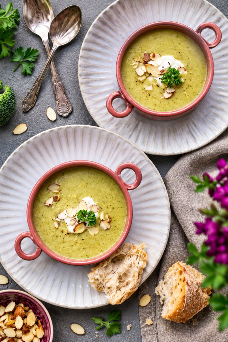 Two bowls of creamy broccoli soup topped with sliced almonds, parsley and a swirl of cream,