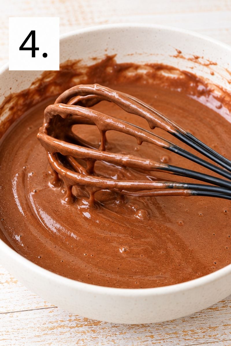 Smooth chocolate crepe batter dripping from a whisk into a mixing bowl.