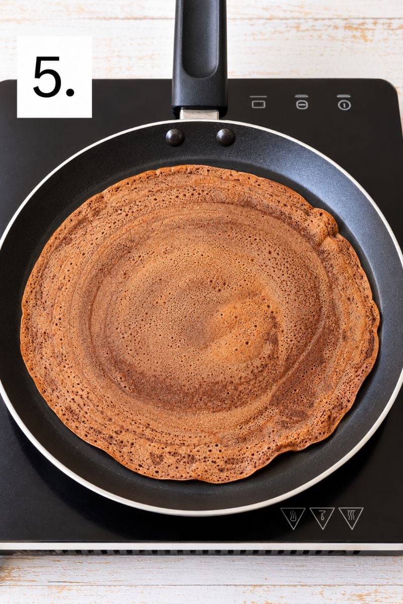 Thin chocolate crepe cooking in a nonstick frying pan.