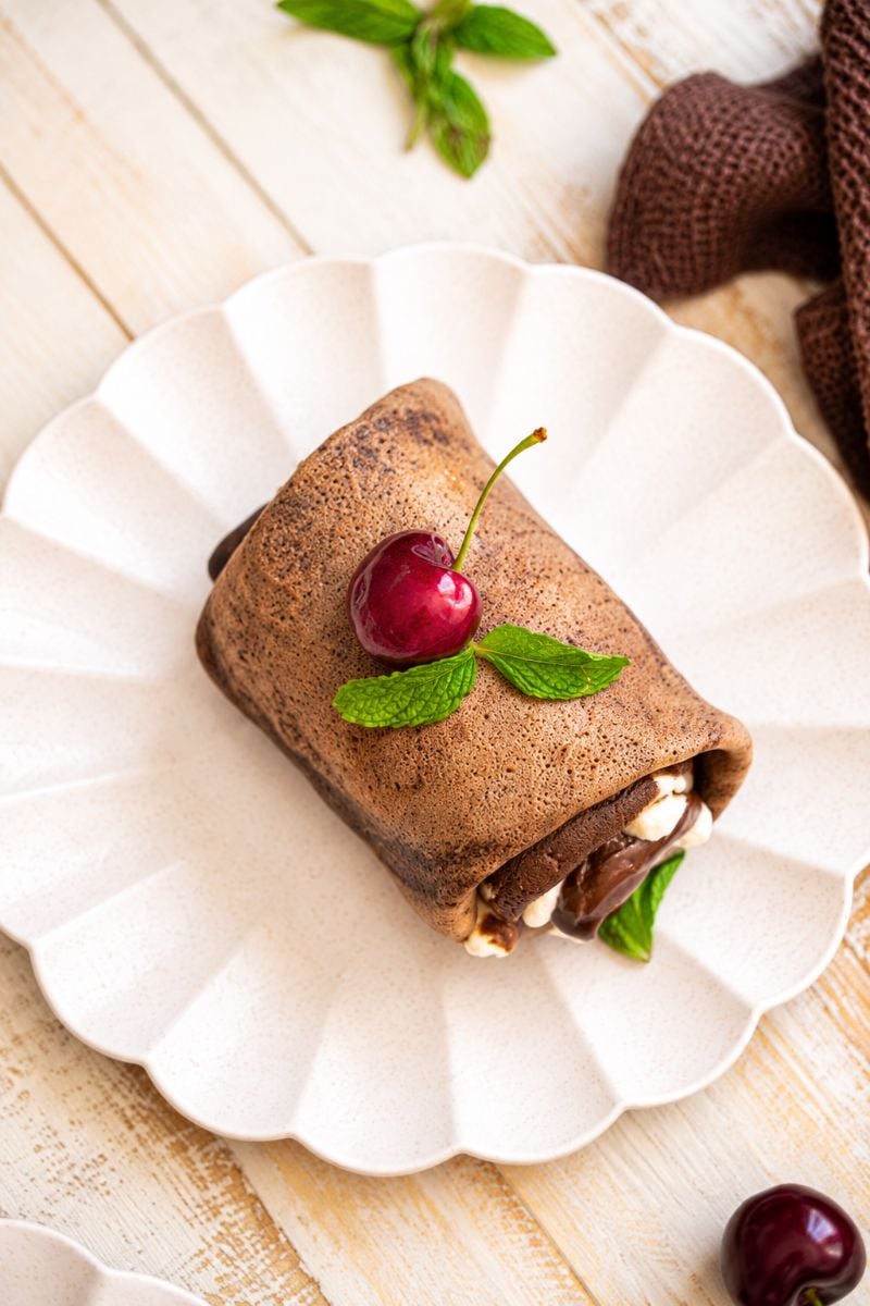 Chocolate crepe roll garnished with a fresh cherry and mint leaves on a white scalloped plate.
