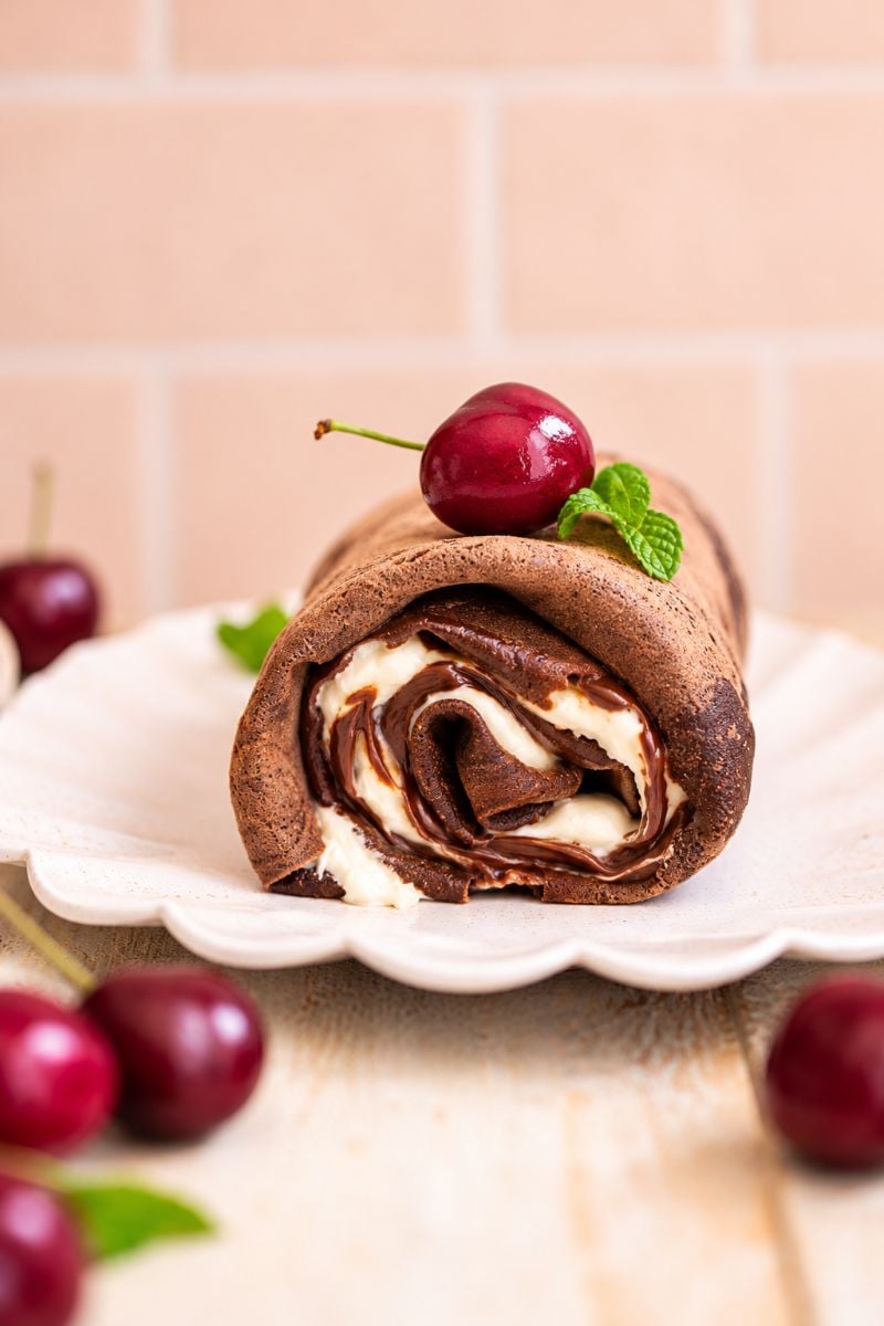 Homemade chocolate crepe roll topped with a cherry and fresh mint on a white plate.
