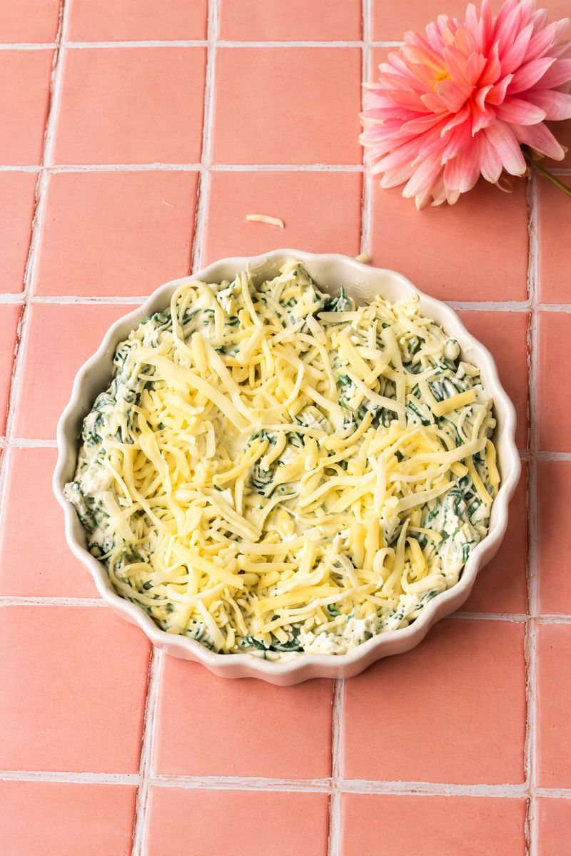 Gluten-free spinach dip in a white baking dish topped with shredded mozzarella cheese before baking.