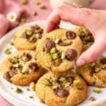 Hand picking up a homemade pistachio chocolate chip cookie from a plate of freshly baked cookies.