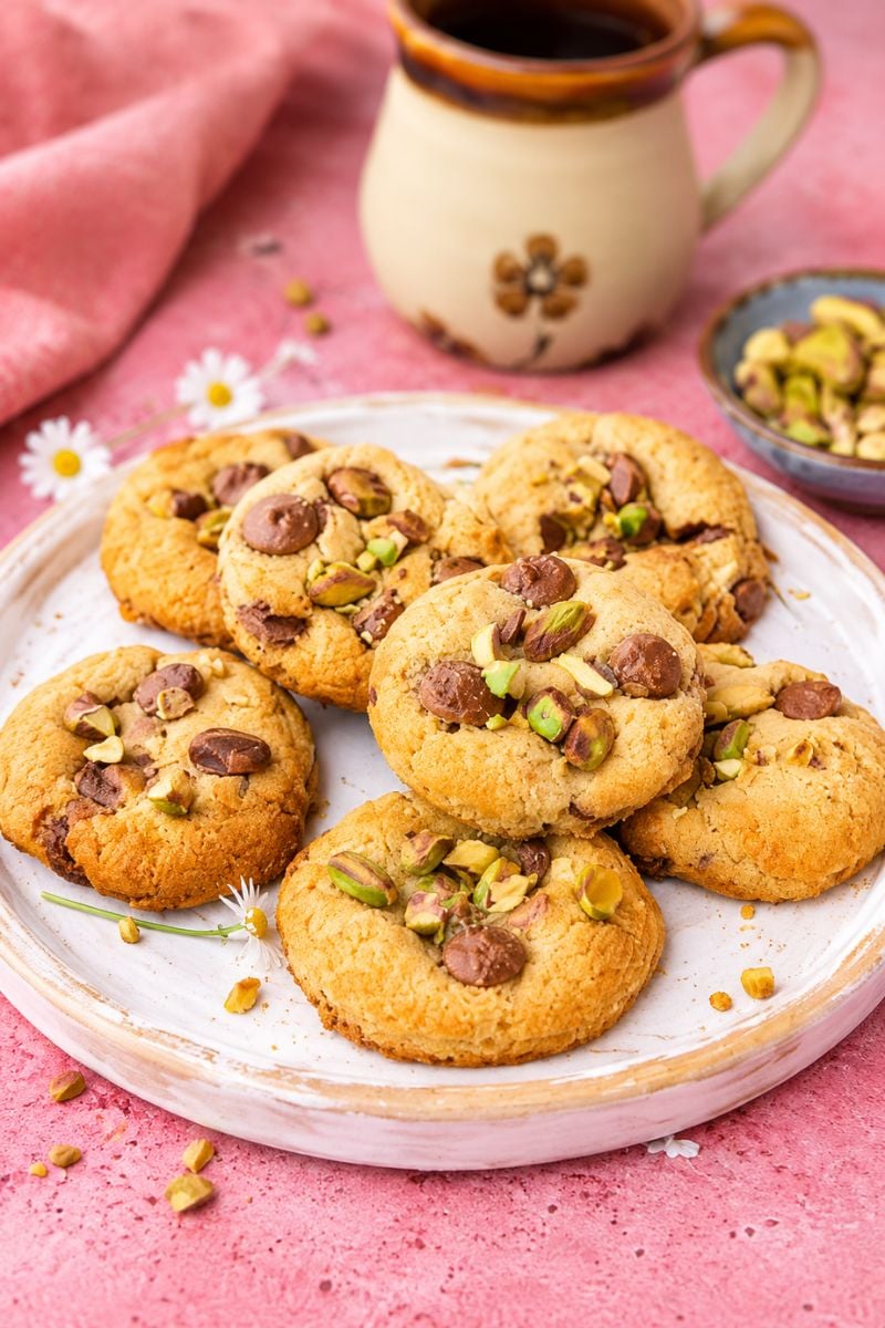 Pistachio chocolate chip cookies arranged on a white plate beside a cup of coffee on a pink background.