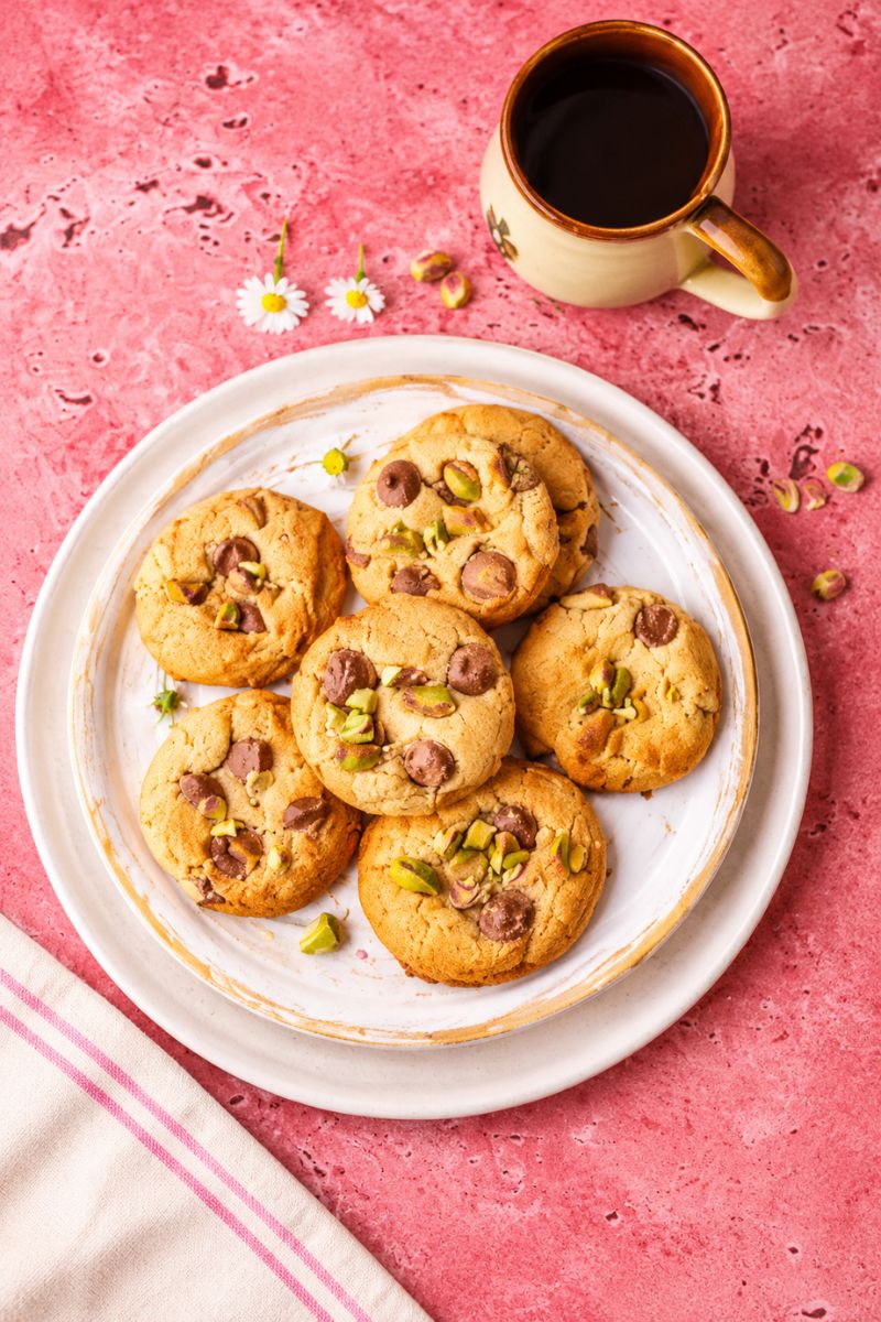 Pistachio chocolate chip cookies arranged on a white plate beside a cup of coffee on a pink background.