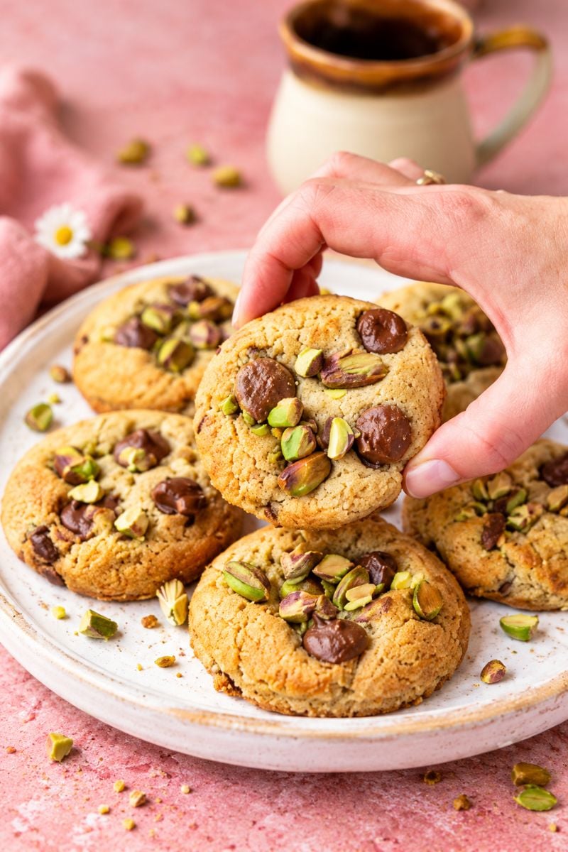 Hand picking up a homemade pistachio chocolate chip cookie from a plate of freshly baked cookies.