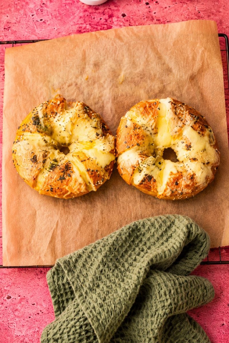 Two golden cream cheese stuffed bagels with melted cheese and herbs resting on baking paper.