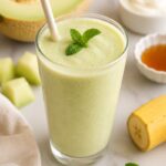 A honeydew smoothie with the ingredients used placed around the smoothie.
