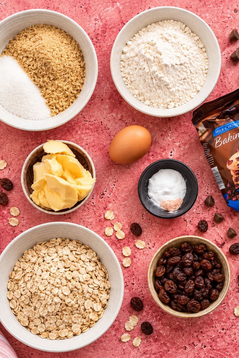 Ingredients for oatmeal raisin chocolate chip cookies arranged in bowls on a pink background.