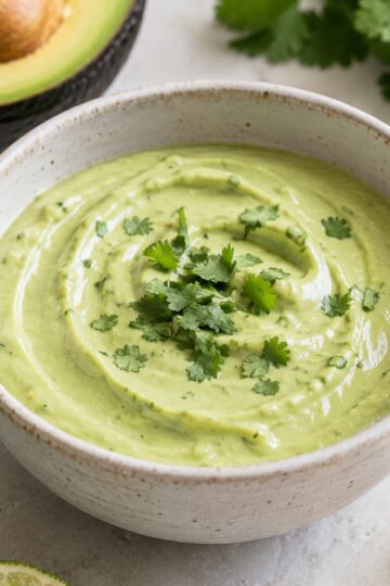 A bowl of avocado lime crema. With avocado and lime next to the bowl.
