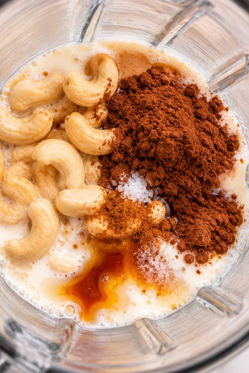 Raw cashews, cocoa powder and maple syrup in a blender ready to make chocolate cashew milk.