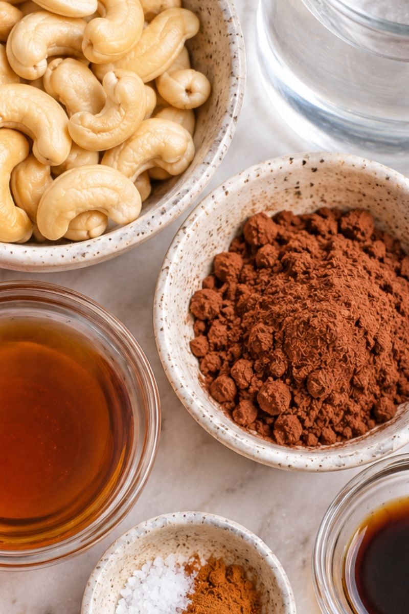 Ingredients for homemade chocolate cashew milk, including raw cashews, cocoa powder, maple syrup and cinnamon.